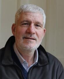 Jim Smith, a white man with white hair and beard, wearing a pale blue shirt and black coat and looking at the camera.