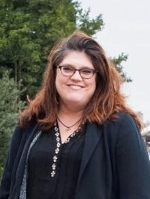 Sandra Squire, a white woman with long brown hair and glasses, wearing a black top and jacket, looking at the camera and smiling with trees in the background