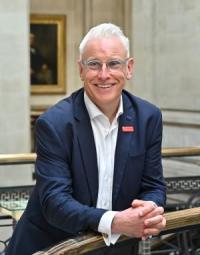 Andy Gutherson, a white man in his fifties, with short white hair and glasses, wearing a dark blue jacket and white shirt, looking slightly away from the camera and smiling