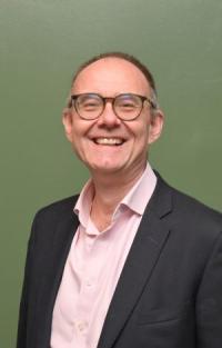 Chris Henning, a white man in his early fifties, with very short receding grey hair and glasses, wearing a dark jacket and pale pink shirt, looking at the camera and smiling