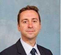 Colin Sykes, a white man in his forties, with short brown hair, wearing a dark suit and tie with a pale shirt, looking at the camera.