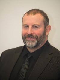 Karl Battersby, a white man in his fifties, very short dark hair with receding hairline and dark/grey beard, wearing a black suit, shirt and tie, looking at the camera and smiling.