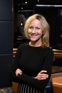 Katie Stewart, a white woman in her forties, with shoulder-length blond hair, wearing a black dress, standing with her arms folded looking at the camera and smiling