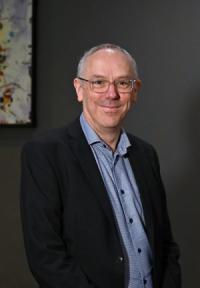 Matt Davey, a white man in his late forties, with very short receding grey hair and glasses, wearing a dark jacket and pale blue shirt, looking at the camera and smiling