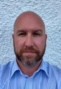 Rhodri Llwyd, a white man, bald with red and grey beard, wearing a striped blue shirt, looking at the camera and smiling.