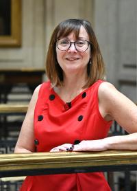Ann Carruthers, a white woman in her early 50s, with shoulder-length mid-brown hair, glasses, wearing a sleeveless red top with black button, smiling at the camera